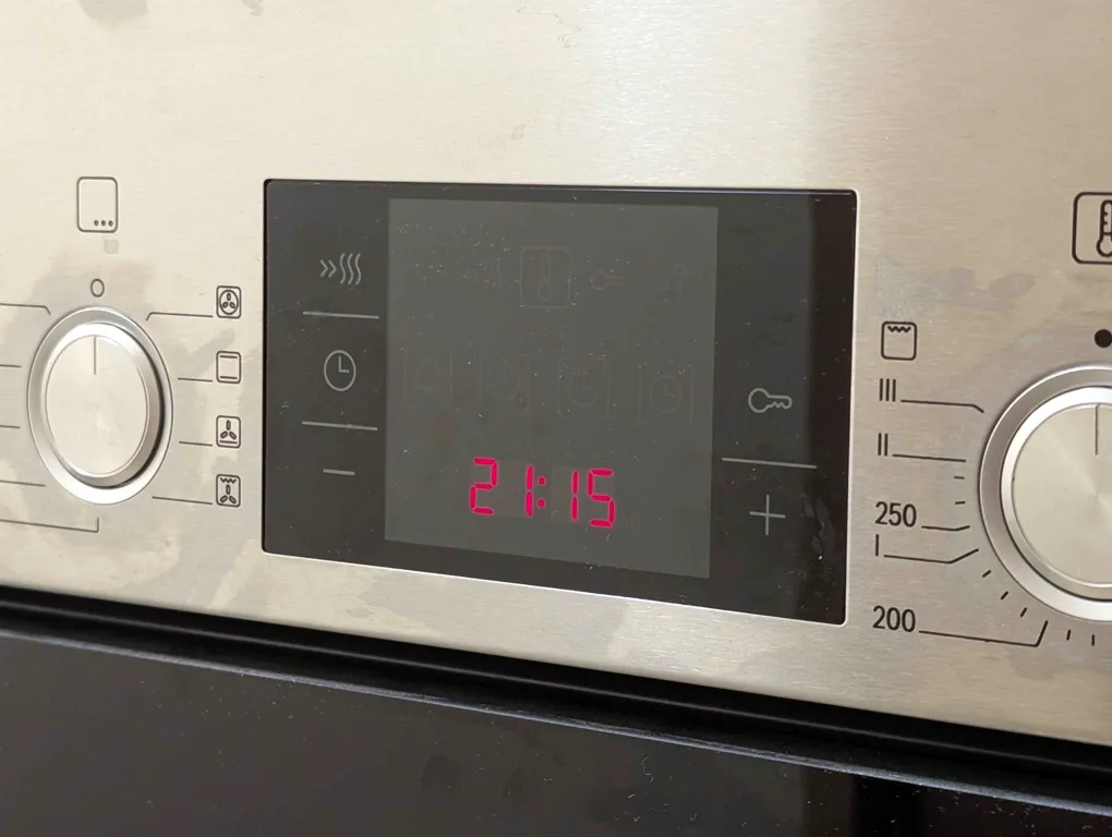 close-up of the oven in my kitchen. from the left: there’s a function knob, the timer panel, and a temperature knob. the timer panel is a touch panel with five buttons, labelled with icons representing heat, a clock, a key, a minus, and a plus. the hour is 21:15.